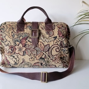 May include: A rectangular handbag with a tapestry floral design in beige, brown, and green. It has brown leather handles, a shoulder strap, and buckle fastenings. The bag has a vintage style and aesthetic.