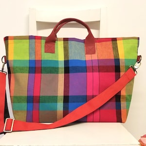 May include: A large tote bag with a vibrant plaid pattern in shades of green, yellow, pink, purple, and black. The bag has red handles and a matching red adjustable shoulder strap. The bag is designed for carrying items.