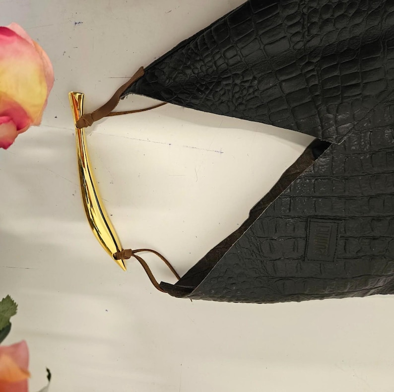 May include: A black leather handbag with a gold-colored handle. The bag has a crocodile skin texture and brown leather straps. The handle is curved and has a shiny finish. The bag is on a white surface.