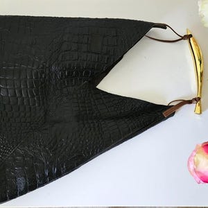 May include: Black clutch with a crocodile texture. The clutch has a gold-colored handle and brown leather straps. The bag is on a white surface with pink and white roses.