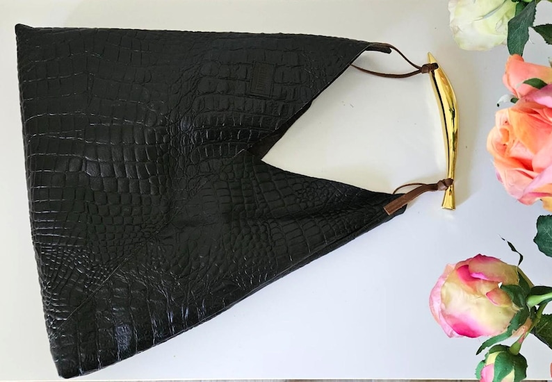 May include: Black clutch with a crocodile texture. The clutch has a gold-colored handle and brown leather straps. The bag is on a white surface with pink and white roses.