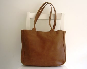 May include: A medium brown leather tote bag with a rectangular shape, featuring two long shoulder straps attached to the top edge with reinforced stitching. The surface of the bag displays a natural, matte texture with subtle variations in color.