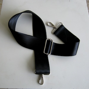 May include: A black adjustable shoulder strap with silver-colored metal hardware. The strap is made of a durable, woven material and features two swivel hooks and a rectangular buckle for length adjustment. Ideal for bags and accessories.