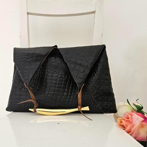 May include: Black clutch with a crocodile texture, featuring a gold-colored bar and brown leather accents. The clutch is folded over and has a triangular shape. The bag is sitting on a white surface.
