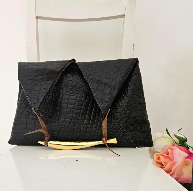 May include: Black clutch with a crocodile texture, featuring a gold-colored bar and brown leather accents. The clutch is folded over and has a triangular shape. The bag is sitting on a white surface.