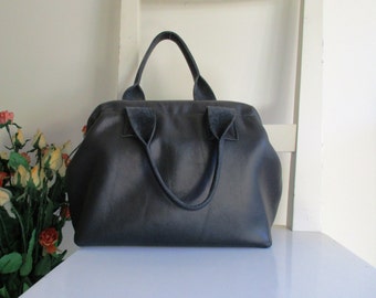 May include: A dark gray leather handbag with two top handles. The bag has a rectangular shape with rounded corners and a structured design. The handles are attached with suede-like material. The bag is sitting on a white surface.
