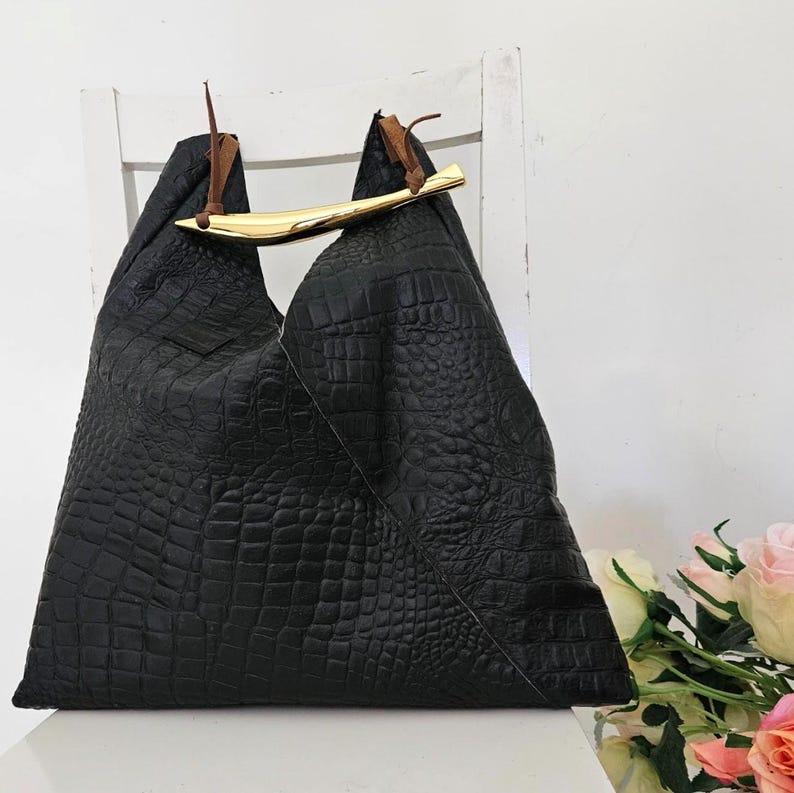 May include: Black leather tote bag with a crocodile texture. The bag features a gold-colored bar handle and brown leather accents. The bag is sitting on a white chair.
