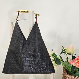 May include: Black leather tote bag with a crocodile texture, hanging from a white chair. The bag has a unique triangular shape and brown leather straps attached to gold-colored handles. A bouquet of roses sits in a wicker basket.