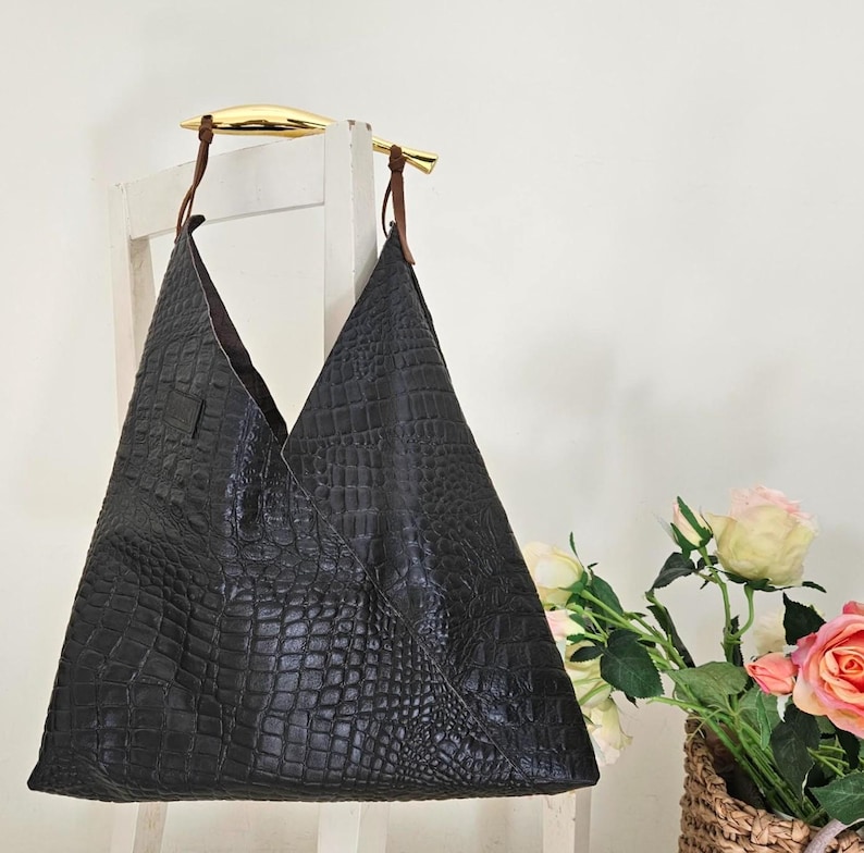 May include: Black leather tote bag with a crocodile texture, hanging from a white chair. The bag has a unique triangular shape and brown leather straps attached to gold-colored handles. A bouquet of roses sits in a wicker basket.