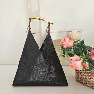 May include: A black, triangular-shaped tote bag with a crocodile texture. The bag has a gold-colored handle and brown leather straps. A bouquet of pink and peach roses sits in a woven basket in the background.