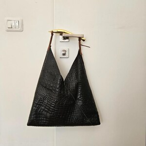 May include: A black leather tote bag with a crocodile skin texture hangs from a door handle. The bag has brown straps and a triangular shape. The door handle is silver and gold, and the door is white.