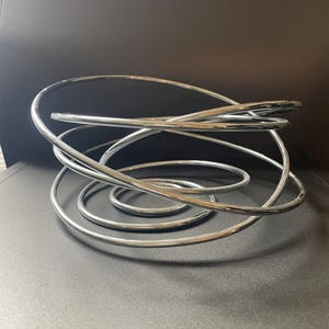 May include: A decorative bowl constructed from a single continuous length of polished chrome metal tubing. The tubing is coiled into a series of overlapping, irregular circular loops that create an open, airy structure with a reflective surface.