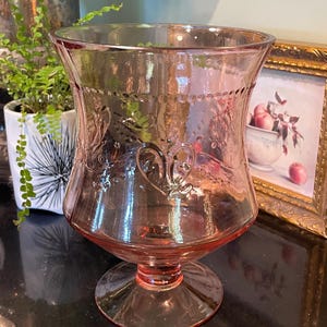 May include: A pink glass vase with a flared top and a decorative pattern. The vase has a round base and a detailed design around the rim. The glass has a slight iridescence, reflecting light. The vase is likely vintage or antique.