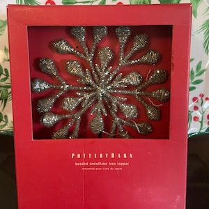 May include: A silver-toned beaded snowflake tree topper displayed inside a red rectangular box. The box features the text Pottery Barn, beaded snowflake tree topper, and ornement pour cime du sapin printed in white on the front panel.