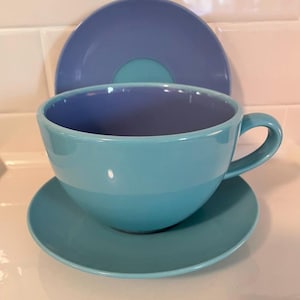 May include: A teal ceramic teacup with a matching saucer and a larger plate in the background. The teacup has a rounded shape with a handle. The saucer and plate are a lighter shade of blue.