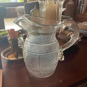 May include: A clear glass pitcher with a large handle and a spout. The pitcher has a textured design with vertical and diamond-shaped patterns. It is sitting on a dark brown surface.