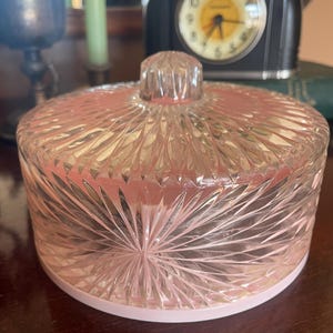 May include: A round, clear glass candy dish with a pink base and a matching lid. The glass features a prominent starburst or sunray pattern radiating from the center of the lid and the sides of the dish, topped with a small, clear glass knob handle.