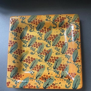 May include: Square ceramic plate with a hand-painted design. The plate features a yellow background with a repeating pattern of green leaves and red berries. Black speckles add detail to the design. The plate is a decorative piece.