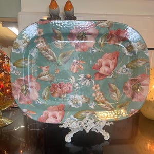 May include: An oval serving platter with a teal background featuring a floral pattern of pink and white flowers with green leaves. The platter has a textured surface and is displayed on a white ornate metal stand. Two small bird figurines sit on top.