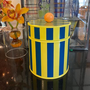 May include: A cylindrical metal canister featuring vertical yellow and dark blue stripes. The lid is topped with a solid orange spherical knob. The canister has a matching striped pattern on the lid that aligns with the body of the container.