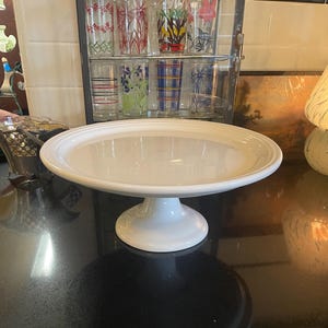 May include: A white ceramic cake stand with a wide, slightly raised rim. The stand has a round base and a central pedestal, creating an elegant display for baked goods. The stand is clean and simple, suitable for various occasions.