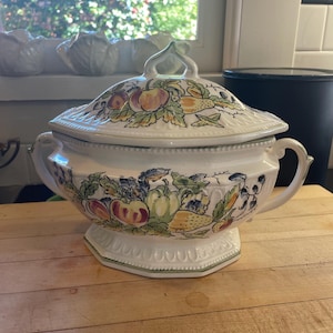 May include: A white ceramic serving dish with a lid, featuring a colorful fruit and vegetable design. The dish has a hexagonal base and two handles. The lid has a loop handle. The dish is on a wooden surface.