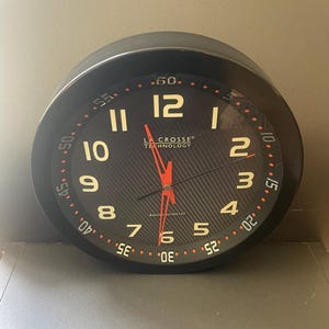 May include: A round black wall clock with a carbon fiber textured face, featuring large off-white Arabic numerals from 1 to 12. The dial includes an outer ring with minute markers, bright red hands, and the text La Crosse Technology Radio Controlled.