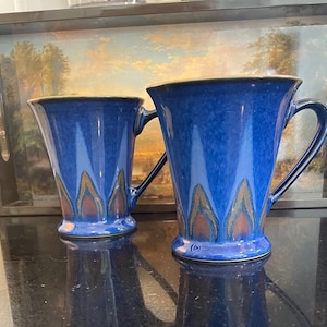 May include: Two blue ceramic mugs with a flared shape and decorative patterns in shades of brown and gold. The mugs have a handle and sit on a reflective black surface. The background features a tray with a landscape painting.