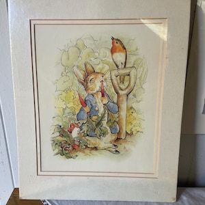 May include: A matted print featuring a watercolor illustration of a rabbit wearing a blue jacket, holding red carrots in a garden. A small bird with an orange breast is perched on the handle of a garden spade next to the rabbit. The artwork is framed in a cream-colored mat board.