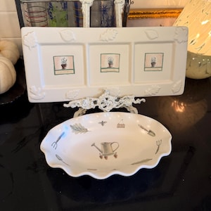 May include: A rectangular off-white ceramic tray with embossed leaf borders and three square panels featuring potted plant illustrations, paired with an oval scalloped-edge dish decorated with garden tool motifs including a watering can.
