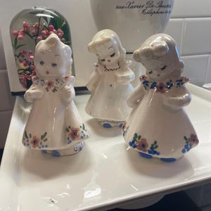 May include: Three vintage porcelain figurines of girls in white dresses with floral accents. The figurines have blue eyes and are arranged on a white tray. A glass dome with pink flowers and a white ceramic cup are in the background.
