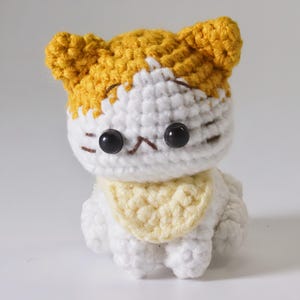May include: A small crocheted cat toy with white and yellow yarn. The cat has black eyes and a yellow bib.