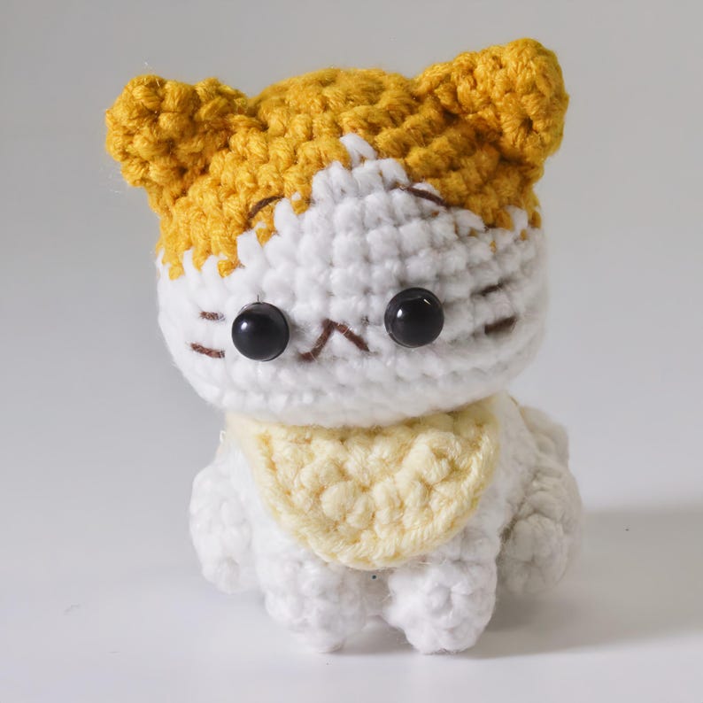 May include: A small crocheted cat toy with white and yellow yarn. The cat has black eyes and a yellow bib.