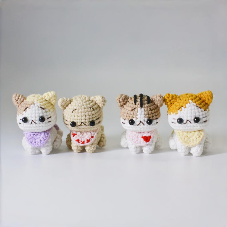 May include: Four crocheted cat plushies with different colored fur and bibs. The cats are all sitting in a row on a white surface.