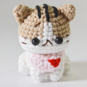 May include: A small crocheted cat wearing a pink sweater with a red heart on the front. The cat has brown and white fur and black eyes.