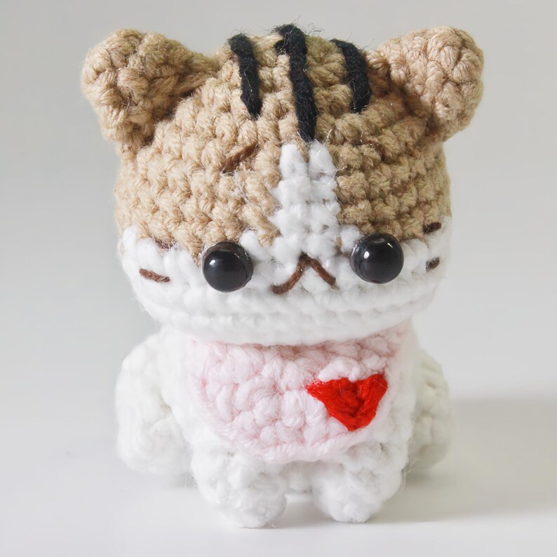 May include: A small crocheted cat wearing a pink sweater with a red heart on the front. The cat has brown and white fur and black eyes.