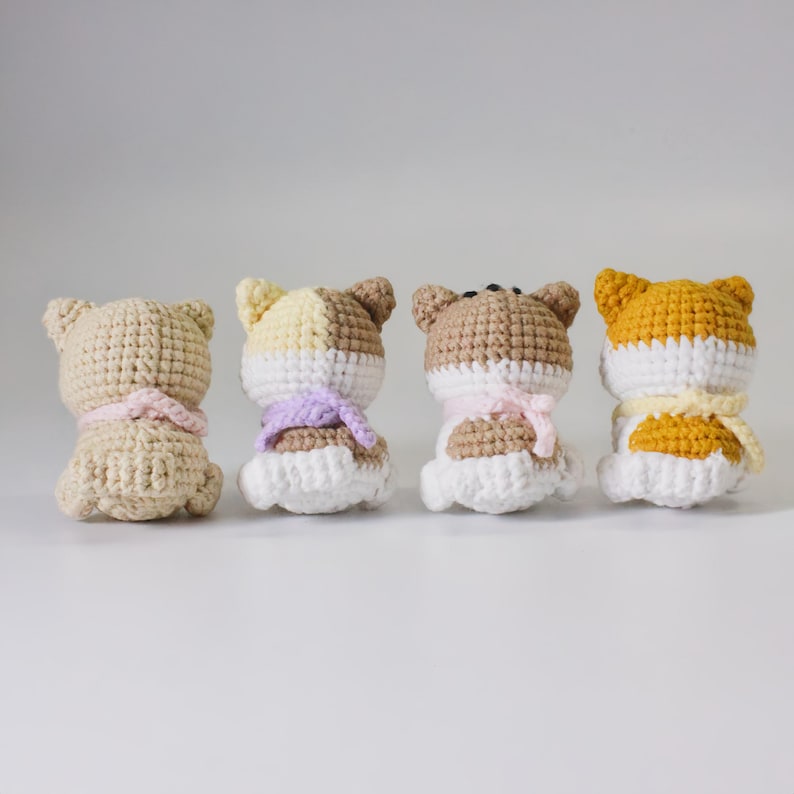 May include: Four crocheted stuffed animals in the shape of dogs. The dogs are all white with different colored patches on their backs. They are wearing crocheted scarves in different colors.