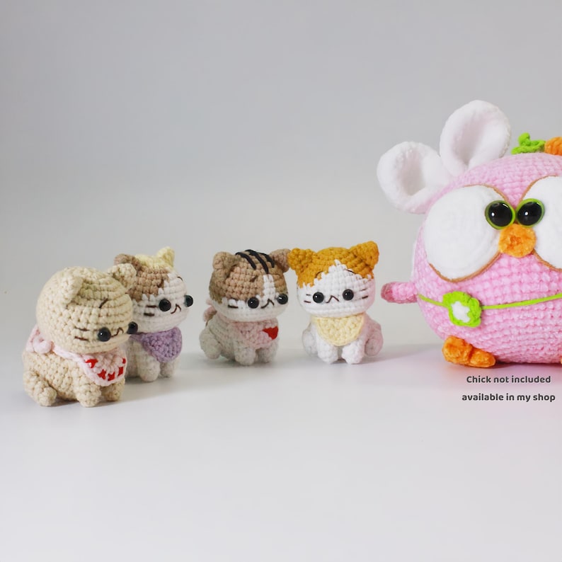 May include: Four crocheted cat plushies in different colors and patterns, sitting on a white surface. The cats are wearing bibs with hearts. A larger crocheted owl plushie is in the background.
