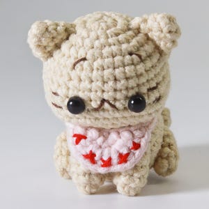 May include: A small crocheted cat toy with a pink bib and red accents. The cat has black eyes and a brown nose.