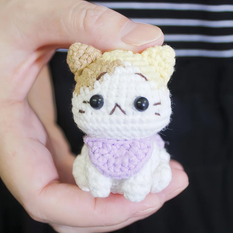 May include: A small crocheted cat toy with white fur, brown and yellow ears, and a purple scarf. The cat has black eyes and a stitched-on black nose and mouth.