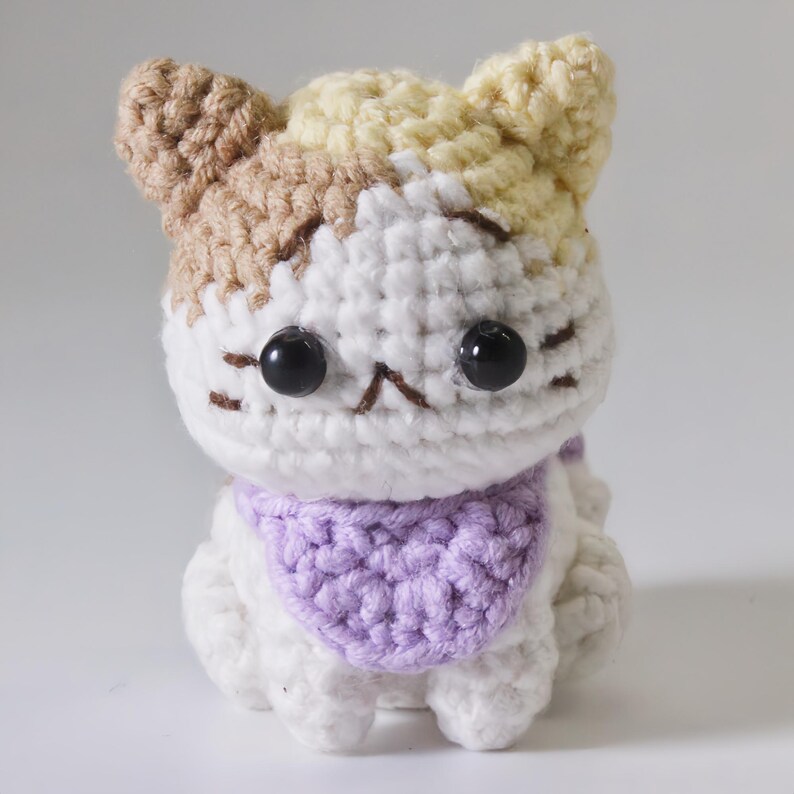 May include: A crocheted cat toy with brown, white, and yellow yarn. The cat has a purple scarf and black eyes.
