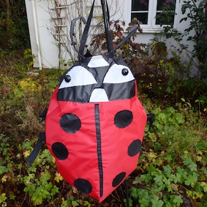 May include: A red ladybird-shaped bag with black spots and a black head. The bag has a drawstring closure and is suspended from a black pole. The background features foliage and a white building.