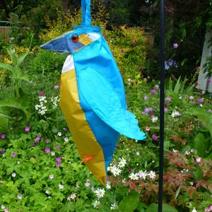 May include: A blue and yellow bird-shaped tote bag with a long handle. The bag is hanging from a metal hook in a garden setting.