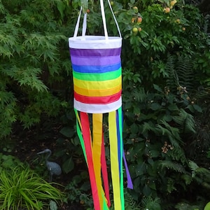 May include: A rainbow-colored windsock with a white top and long streamers hanging down. The windsock is hanging from a tree branch.