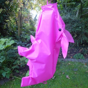 May include: A bright pink, fabric, hanging toy shaped like a rhinoceros with a black eye. The toy is hanging from a string in a grassy area.