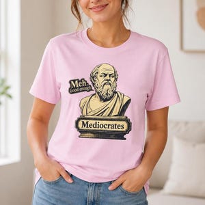 May include: A light pink short-sleeved t-shirt featuring a graphic of a bearded philosopher bust on a pedestal. The text reads "Meh. Good enough" in a speech bubble and "Mediocrates" on the pedestal base. The design uses muted beige and black tones.