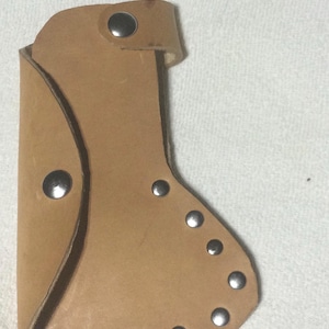 May include: A light brown leather sheath with a curved shape, featuring multiple silver rivets along the edges and a single rivet at the top. The sheath appears to be designed to hold a tool.