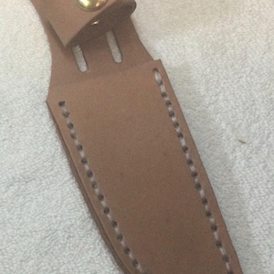 May include: A light brown leather knife sheath with a rounded top and a strap secured by a gold-colored button. The sheath is stitched with white thread along the edges.