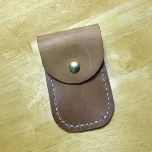 May include: A small, brown leather pouch with a rounded flap secured by a brass snap. The edges are stitched with white thread. The pouch is rectangular with a curved top and is set against a wooden surface.