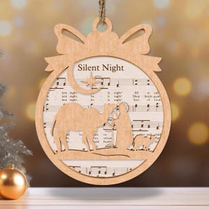 May include: A wooden Christmas ornament shaped like a circle with a bow at the top. The ornament features a cutout of the nativity scene with a star and a crescent moon. The ornament is decorated with sheet music for the song "Silent Night".